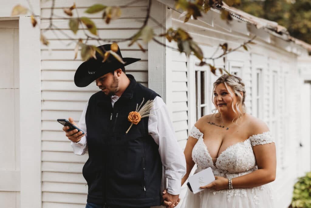 Wedding photography capturing a bride in an off-shoulder gown and a cowboy groom checking his phone, against a backdrop of a white vintage house in Oklahoma.