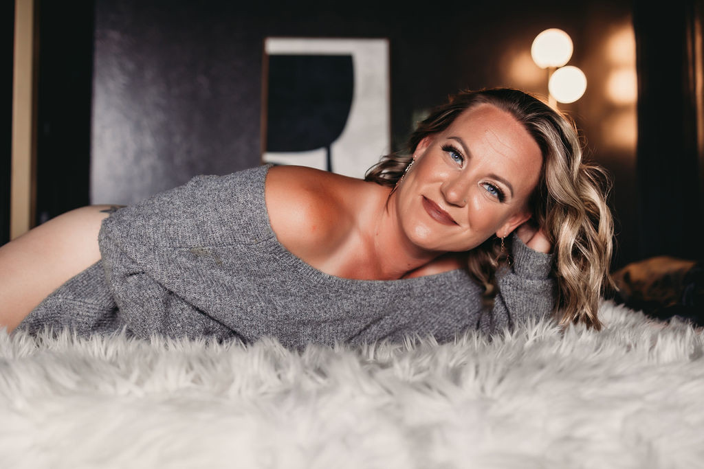 Smiling woman lying on a bed in a cozy off-shoulder sweater during a boudoir experience