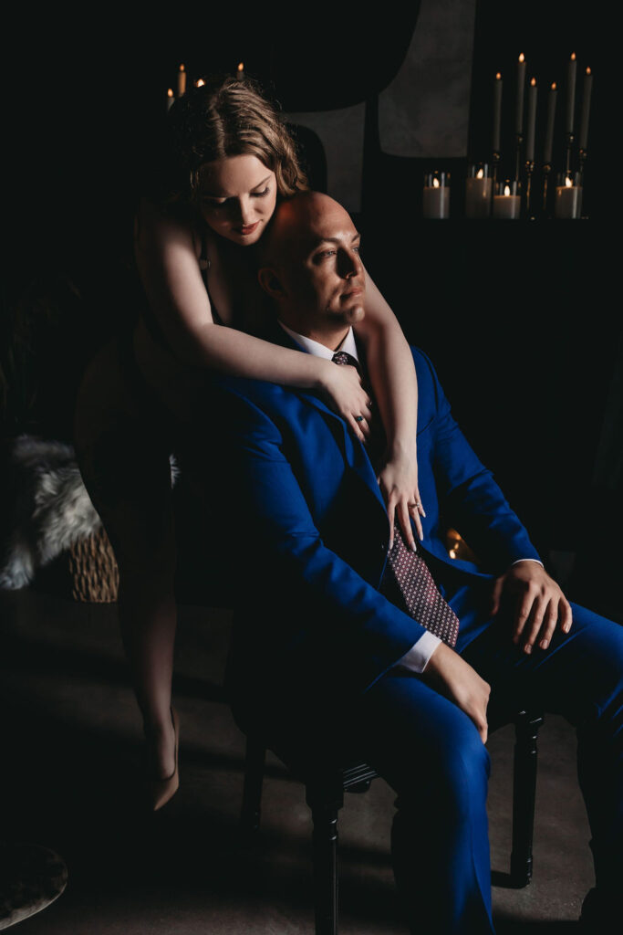 A woman embraces her partner from behind as he sits in a chair, exemplifying the intimacy captured in couples boudoir photography.