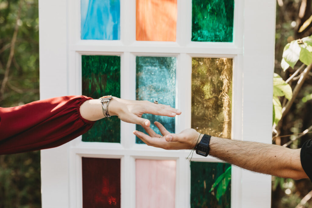 A close-up of a couple's hands reaching toward each other in front of a vibrant stained glass backdrop, captured by a wedding photographer.