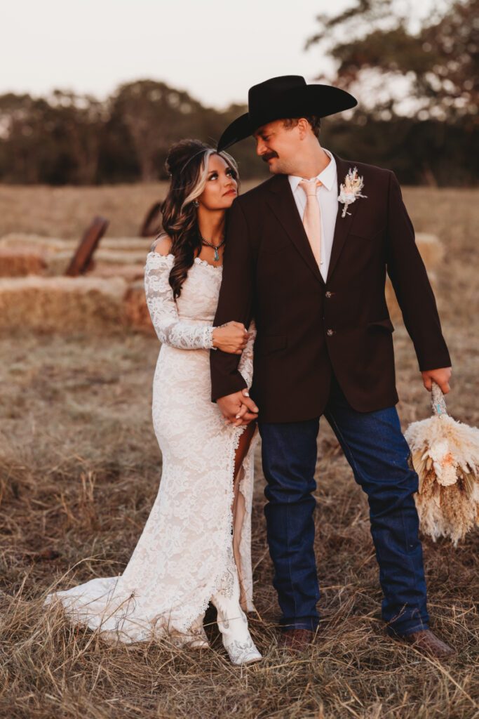 A bride in a lace gown and cowboy boots stands lovingly beside her groom in a cowboy hat, captured by a wedding photographer in a rustic field setting.