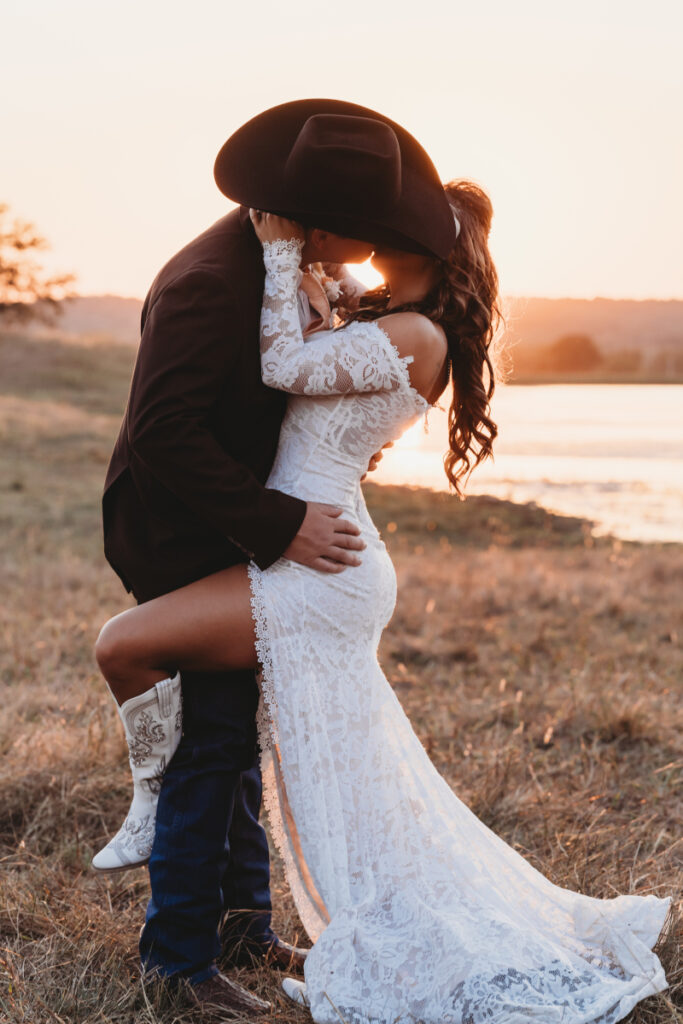 A bride in a lace gown and cowboy boots shares a romantic sunset kiss with her groom in a cowboy hat, captured by a wedding photographer in a field.