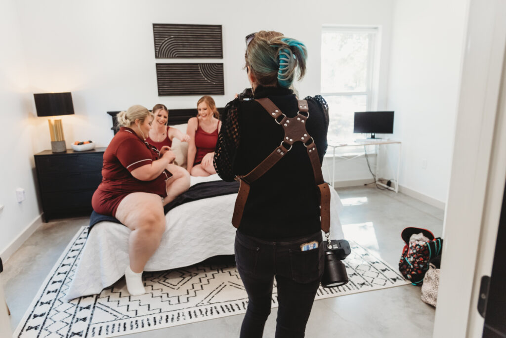 A wedding photographer with a leather camera harness captures candid moments of a bride and bridesmaids laughing together in matching pajamas before the ceremony.