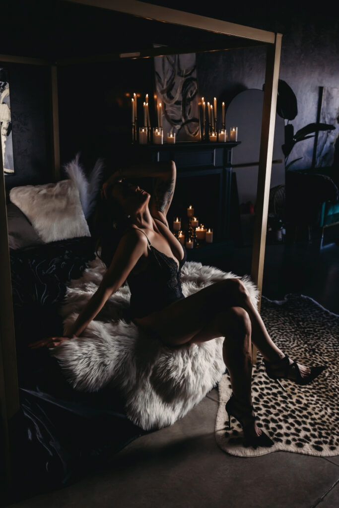 Woman in lingerie seated on the edge of the bed with candles in the background – boudoir photography.